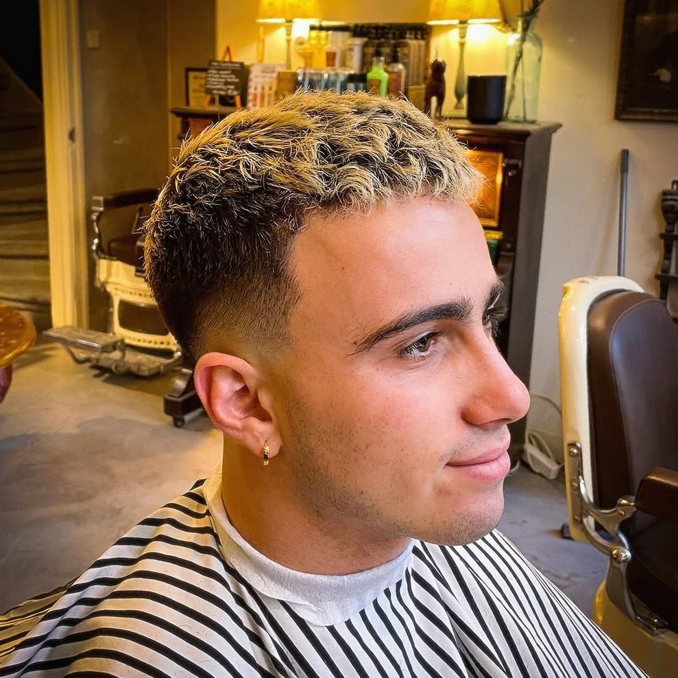 Skin fade met geblondeerde krullende punten, klant in profiel in een gestreepte kapmantel