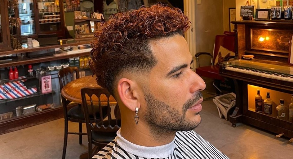 Klant in een gestreepte kapmantel met krullen bovenop en een skin fade aan de zijkant, in de salon met de piano op de achtergrond